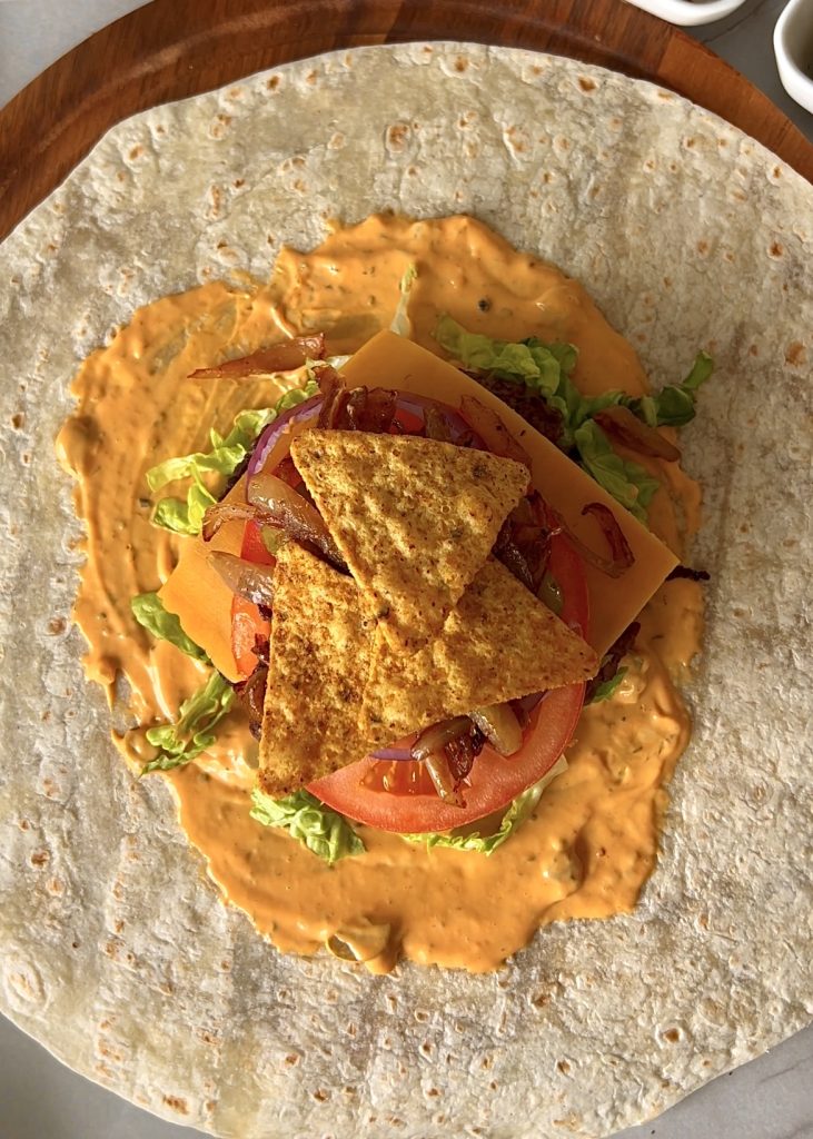 Assemble the Wrap
Lay a tortilla flat and spread sauce in the center. Layer shredded lettuce, cooked beef, cheese, tomato slices, red onion, caramelized onions, pickle slices, and crushed chili nacho chips for crunch.