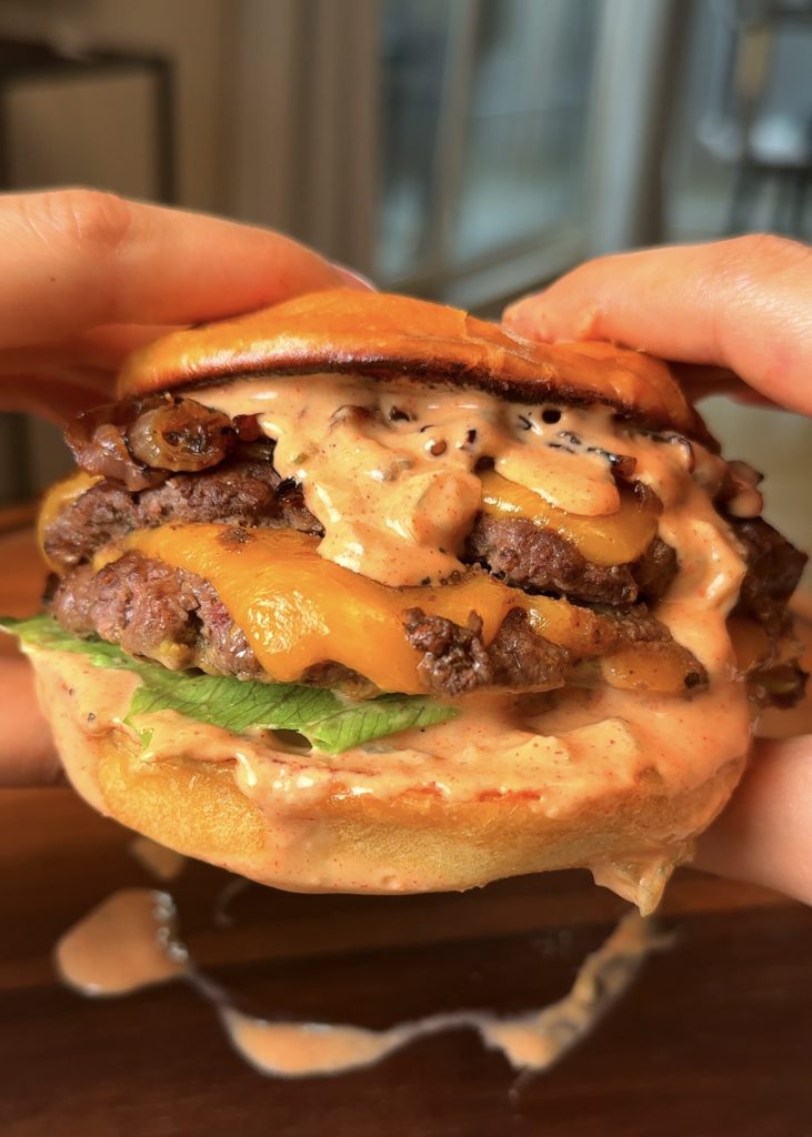 Assemble the burgers by spreading sauce on the bun, adding tomato and lettuce, stacking two cheesy patties, adding caramelized onions, more sauce, and finishing with the top bun.